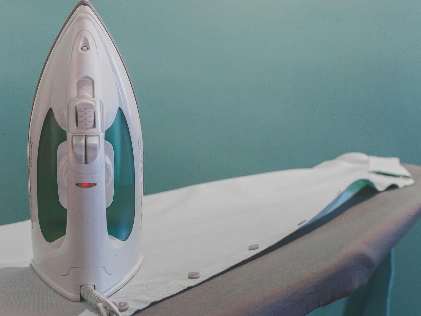 white and teal steam clothes iron plugged on ironing board